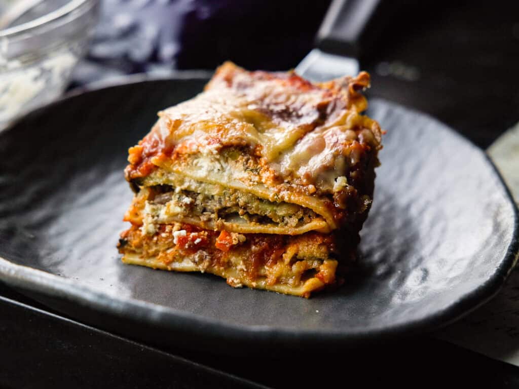 A slice of lasagna with layers of pasta, eggplant, cheese, and sauce sits on a dark plate. The lasagna is golden and slightly crispy on top, with a rich, savory filling visible between the layers.